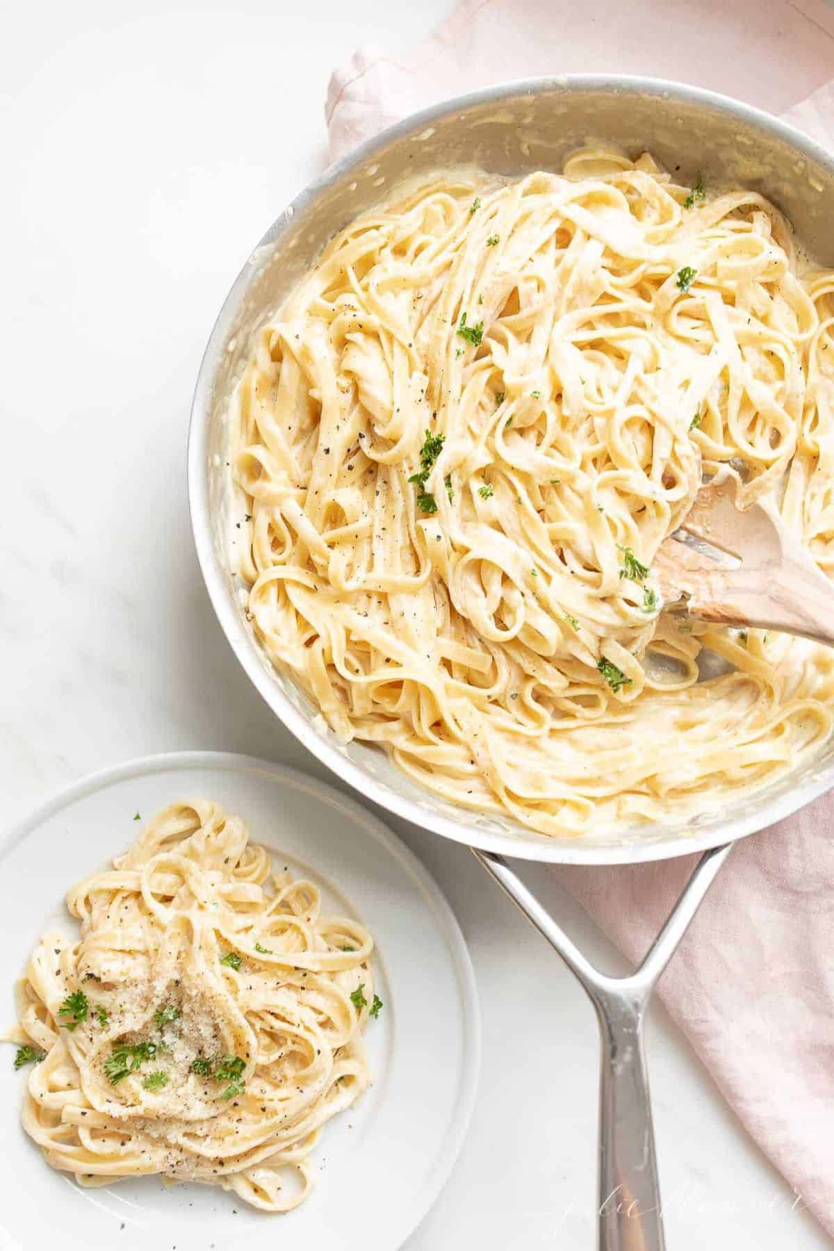 Fettuccine Alfredo in a metal pan on a white surface with a pink linen napkin to the side.