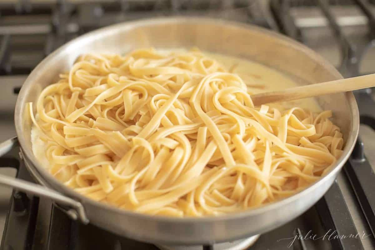 Fettuccine Alfredo in a metal pan with parmesan cheese being mixed in. 