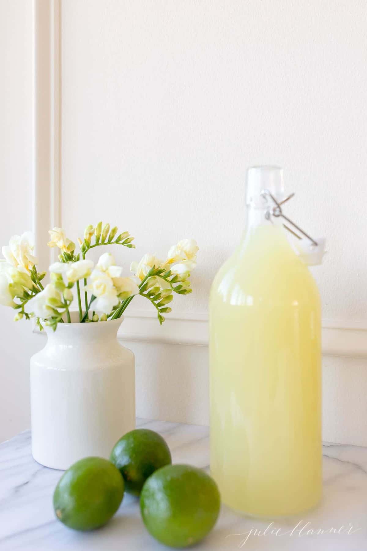 A yellow juice liquid stored in a glass carafe; a vase full of yellow flowers sits to the side. 