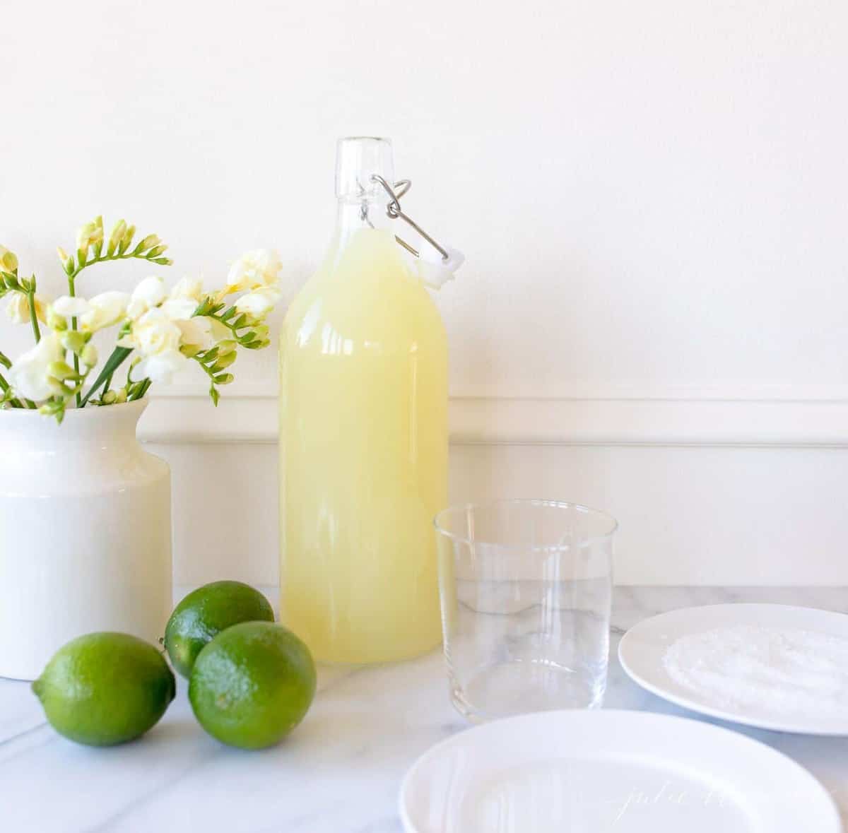 Homemade margarita mix in a glass carafe; a vase full of yellow flowers sits to the side. 