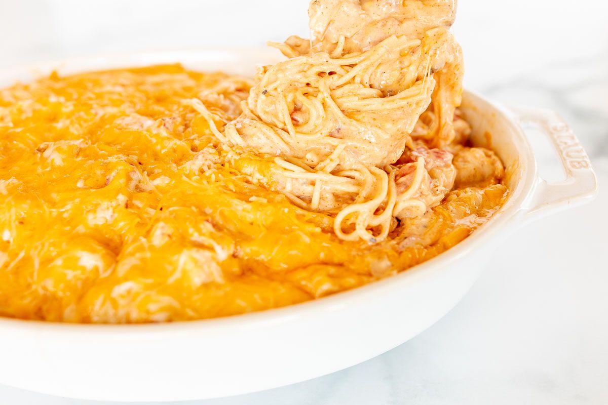 Mexican spaghetti in an oval baking dish, topped with shredded cheese, a scoop being pulled out with a spoon.