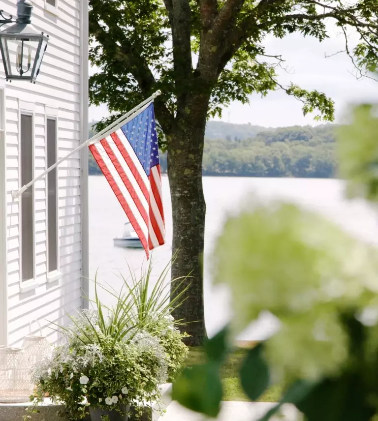 An American flag is mounted on the side of a white house near a lake, with trees and greenery visible.
