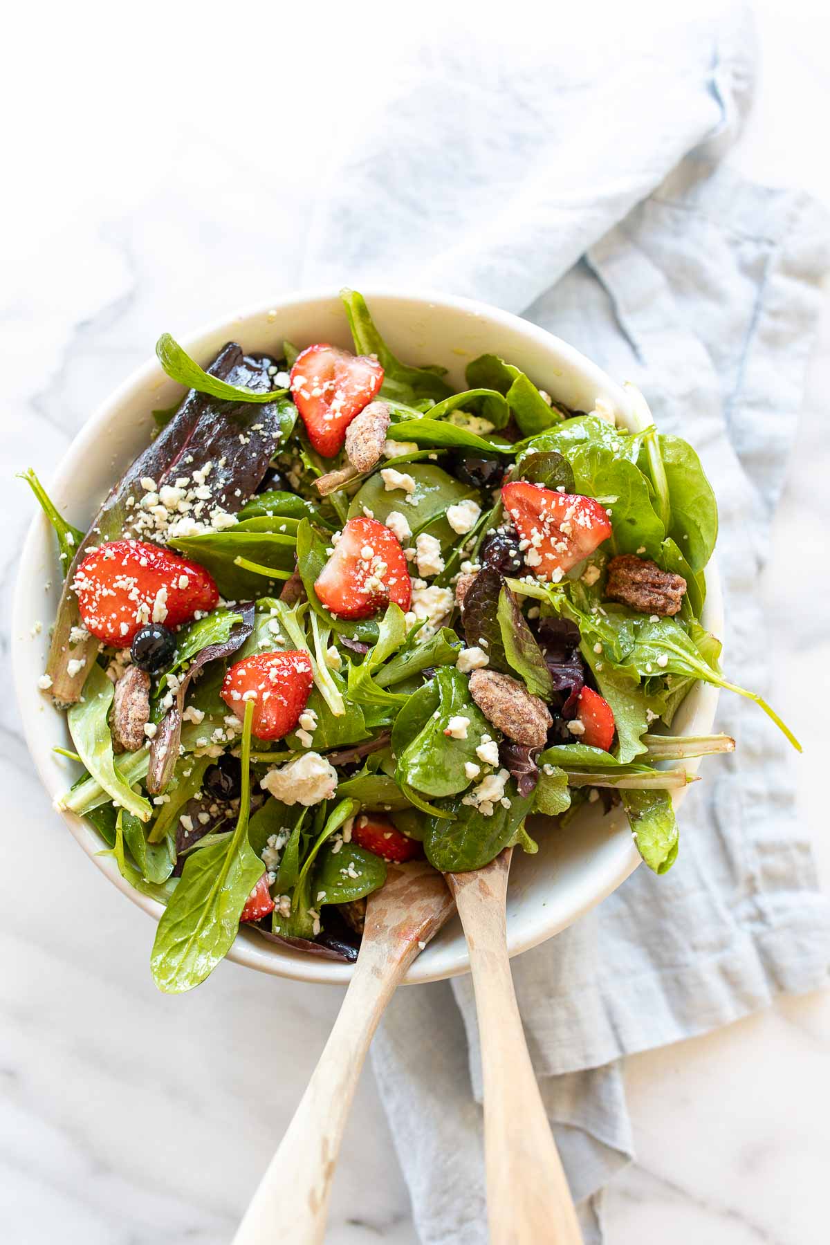 A fresh spring salad with berries, crumbled cheese and nuts in a bowl with wooden serving utensils.