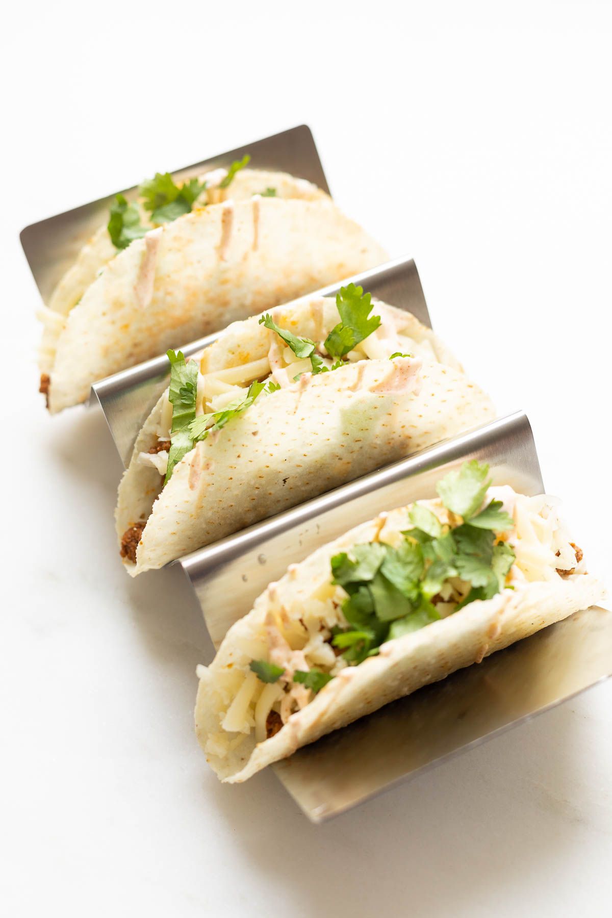 A silver taco holder, holding tacos stuffed with a beef taco meat recipe and topped with cheese and lettuce.