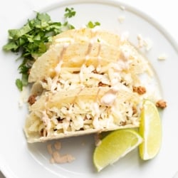 Beef tacos on a white plate, garnished with cheese, sour cream, and limes and cilantro on the plate.