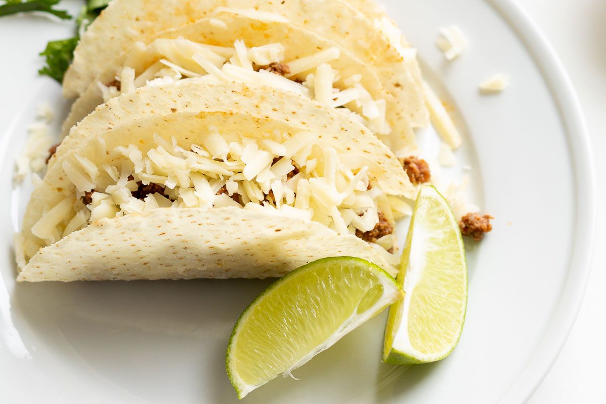 Beef tacos on a white plate, garnished with cheese, sour cream, and limes and cilantro on the plate.