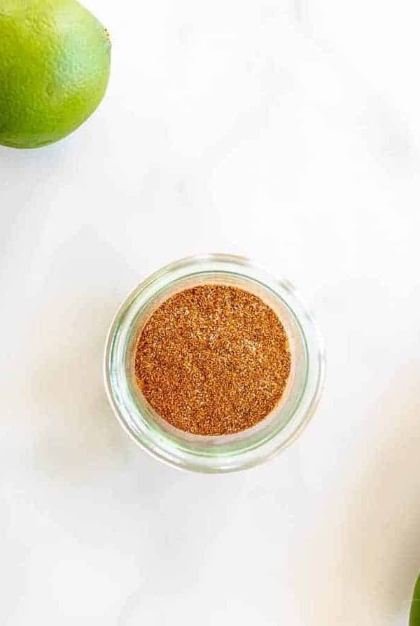 Marble surface with a clear jar full of fajita seasoning, limes and jalapeno to the side.