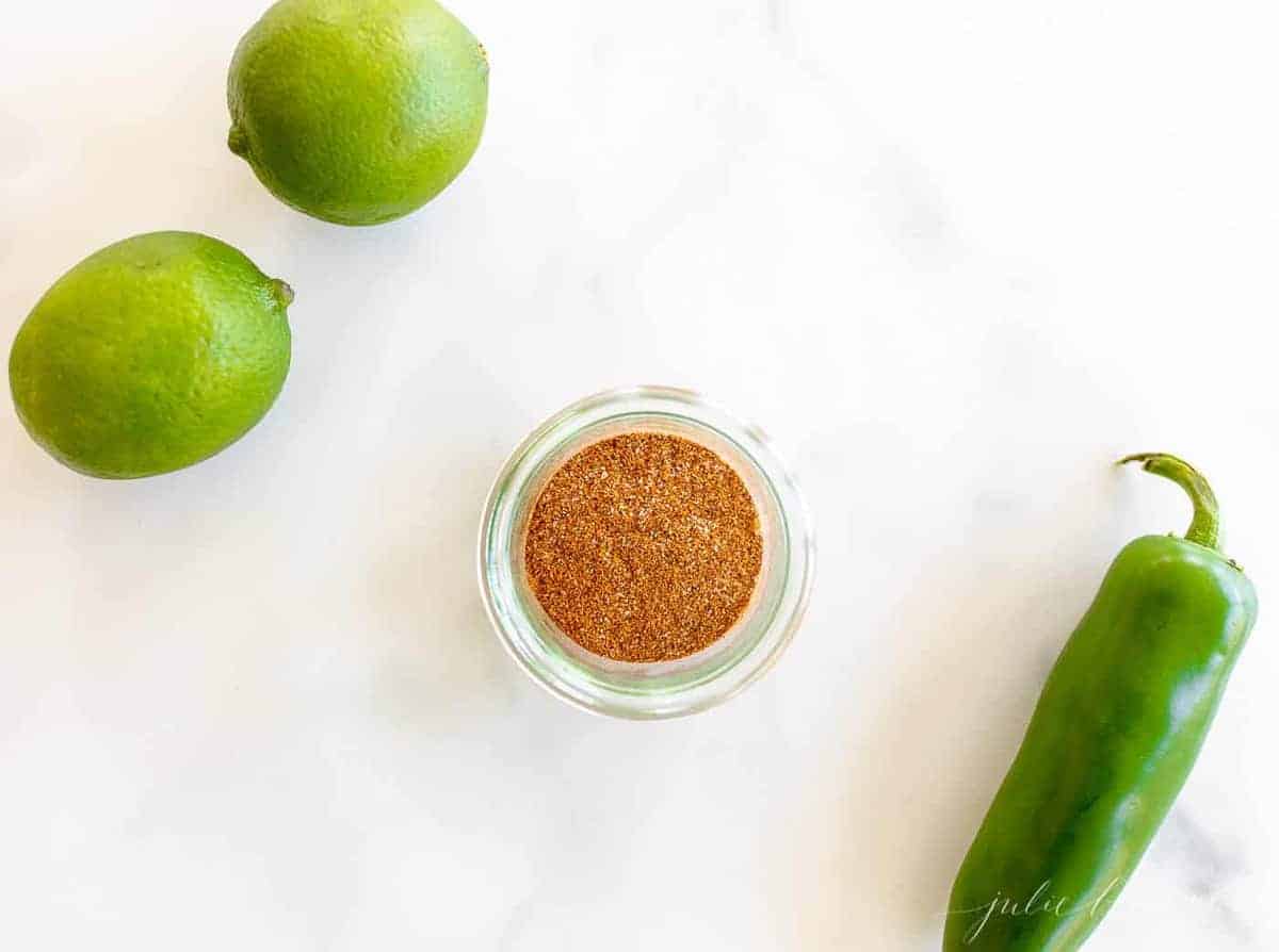 Marble surface with a clear jar full of fajita seasoning, limes and jalapeno to the side.