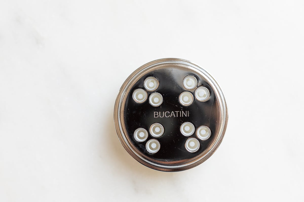 Top view of a black bucatini noodles maker disc with small circular holes on a marble surface.