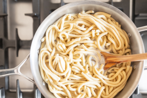 A pot of cooked bucatini pasta with a wooden spoon stirring it on a stove.