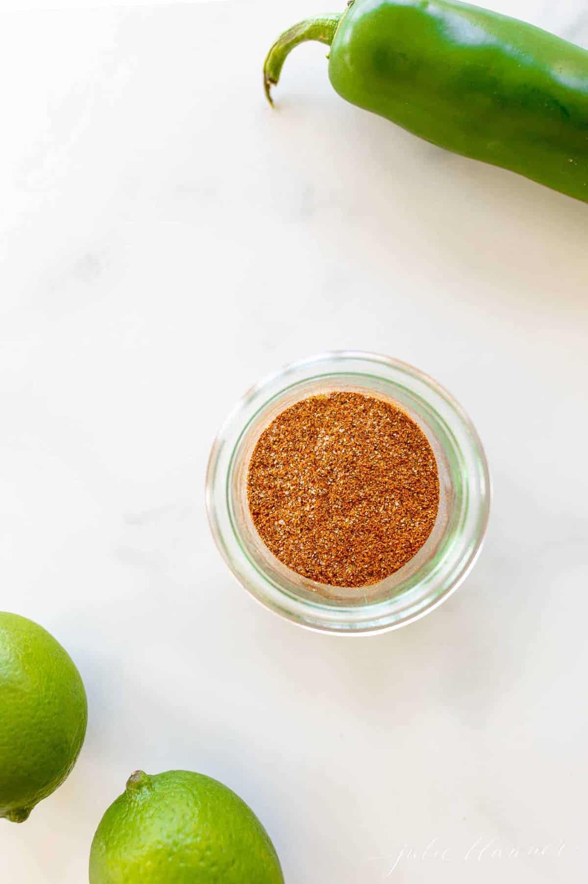 Marble surface with a clear jar full of fajita seasoning, limes and jalapeno to the side.