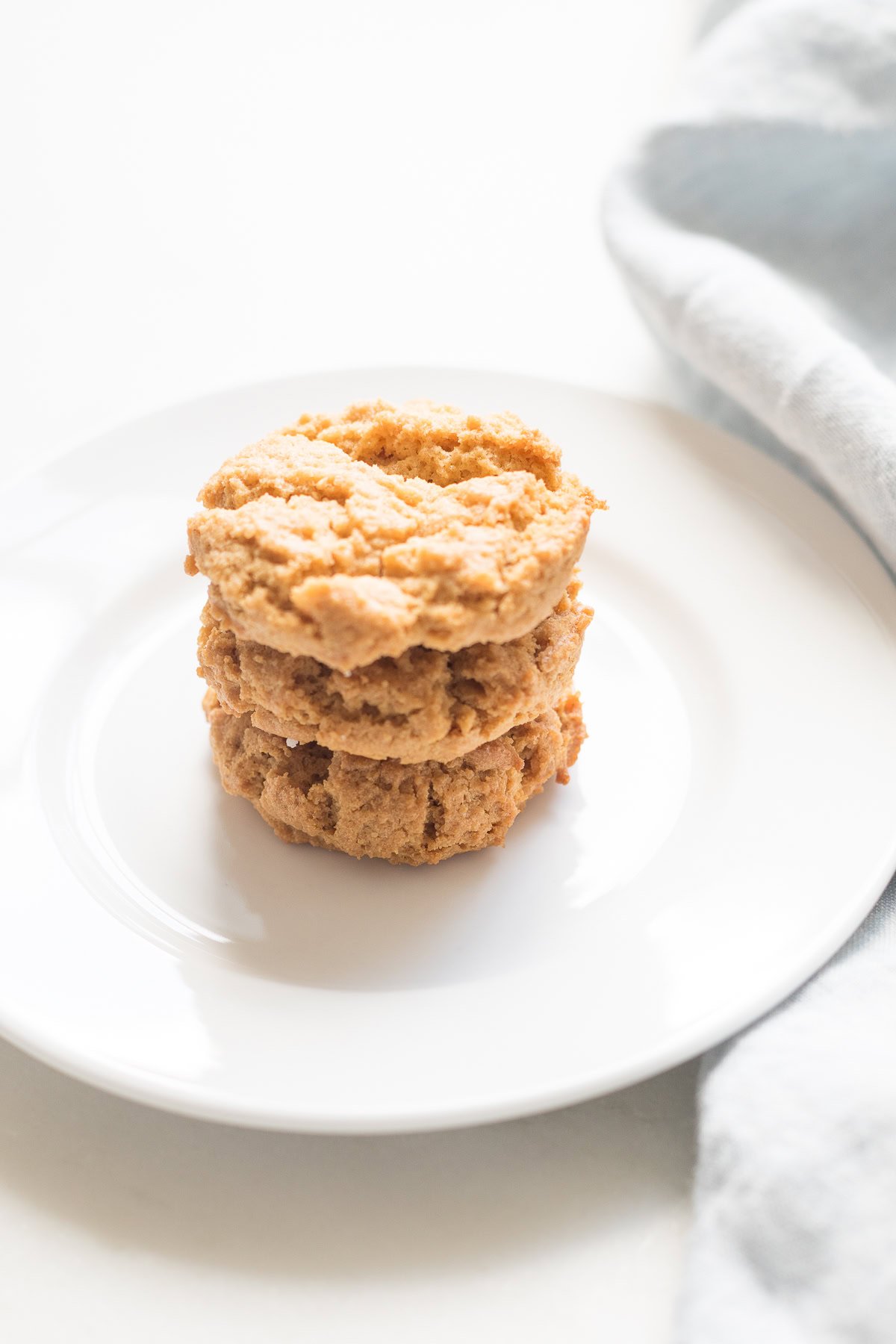 Three flourless peanut butter cookies stacked on a white plate beside a light gray cloth, offering a delightful treat that's both simple and healthy.