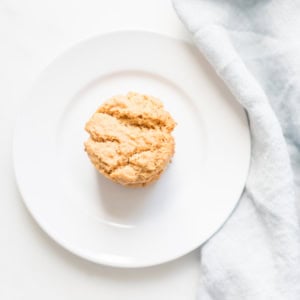 A single muffin rests on a white plate beside a light gray cloth, reminiscent of the simplicity of flourless peanut butter cookies.