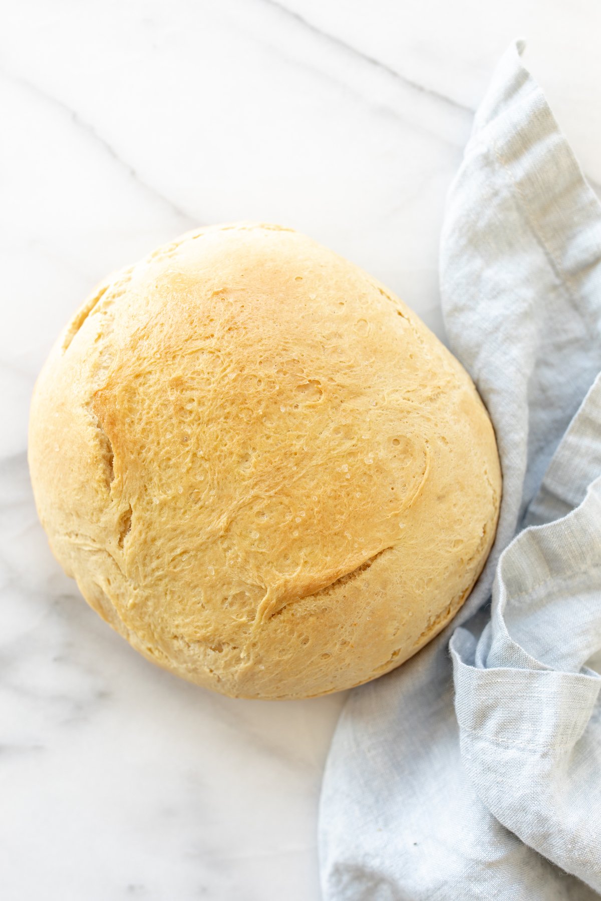 A marble surface with a round loaf of Dutch oven homemade bread, blue linen napkin to the side.