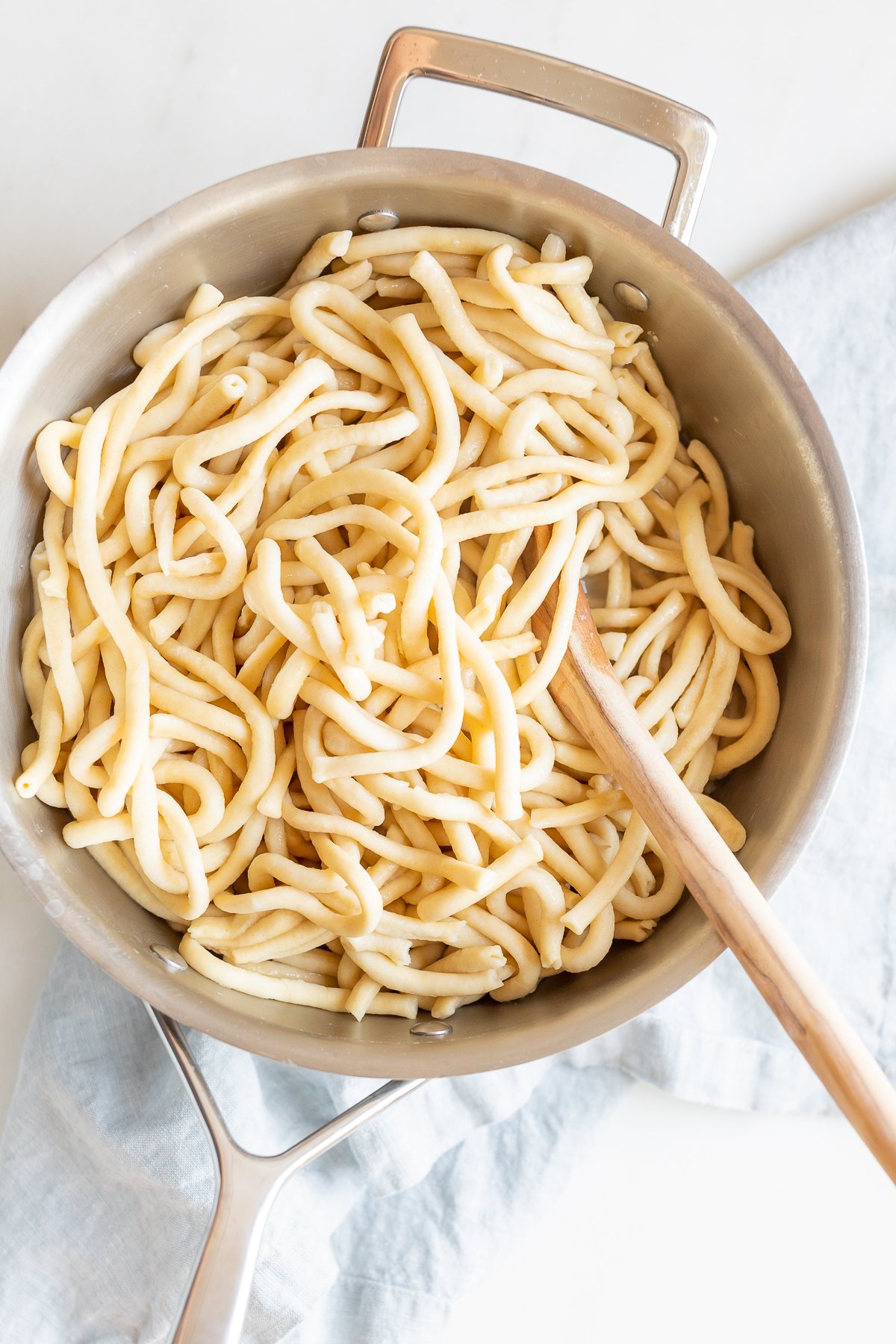 A stainless steel pot filled with cooked bucatini noodles and a wooden spoon, placed on a white surface.