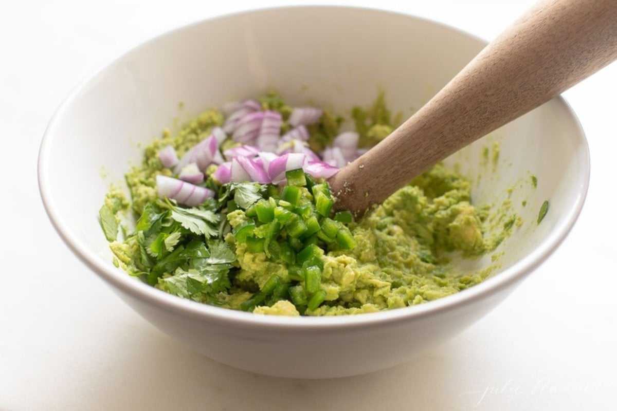 A white bowl full of the beginning of a homemade guacamole recipe, with a wooden pestle.