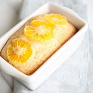 A quick orange bread in the form of a rectangular loaf cake topped with mandarin orange slices, baked in a white ceramic dish, placed on a light gray cloth.