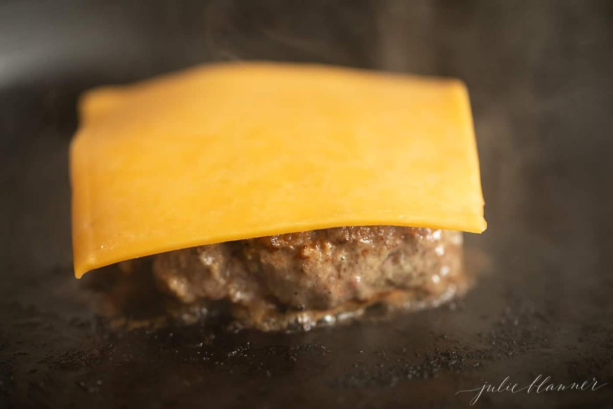 A burger on the stove in a cast iron pan, slice of cheese on top before it melts.