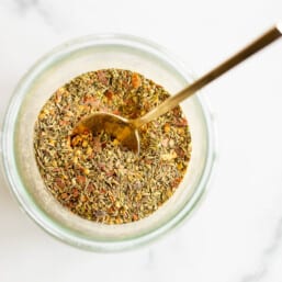 A clear glass jar of italian seasoning blend, gold spoon sticking out.