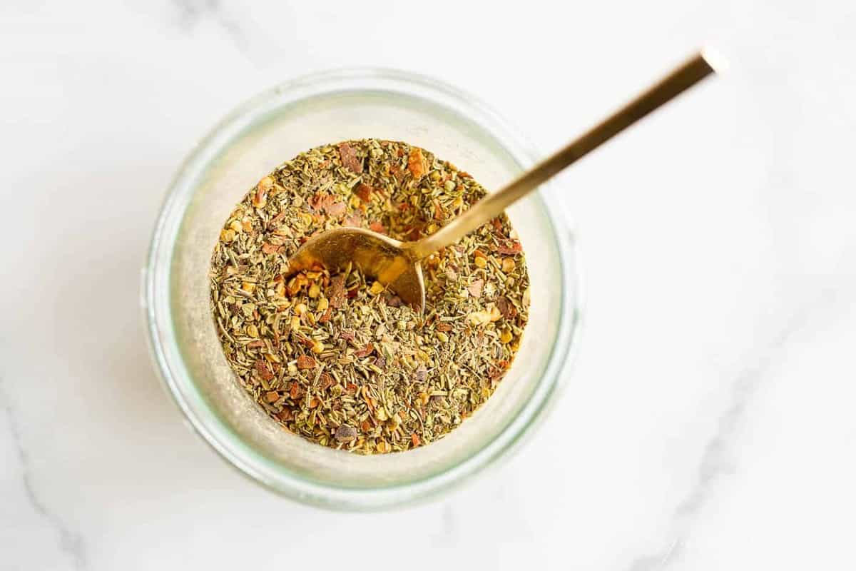 Homemade Italian seasoning in a glass bowl on a marble surface. A gold spoon is inside. 