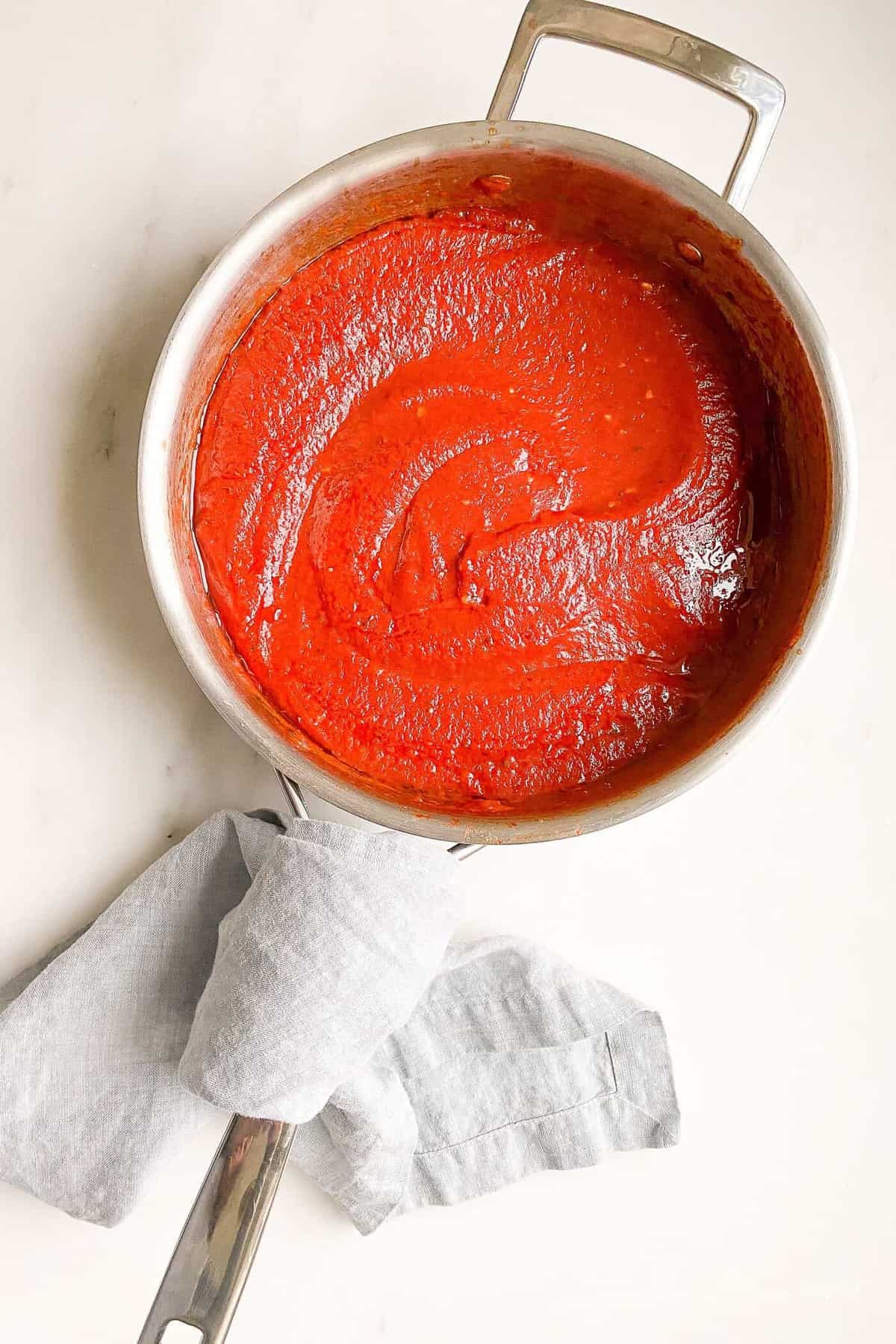 A silver sauce pan filled with homemade pizza sauce, blue napkin wrapped around handle.