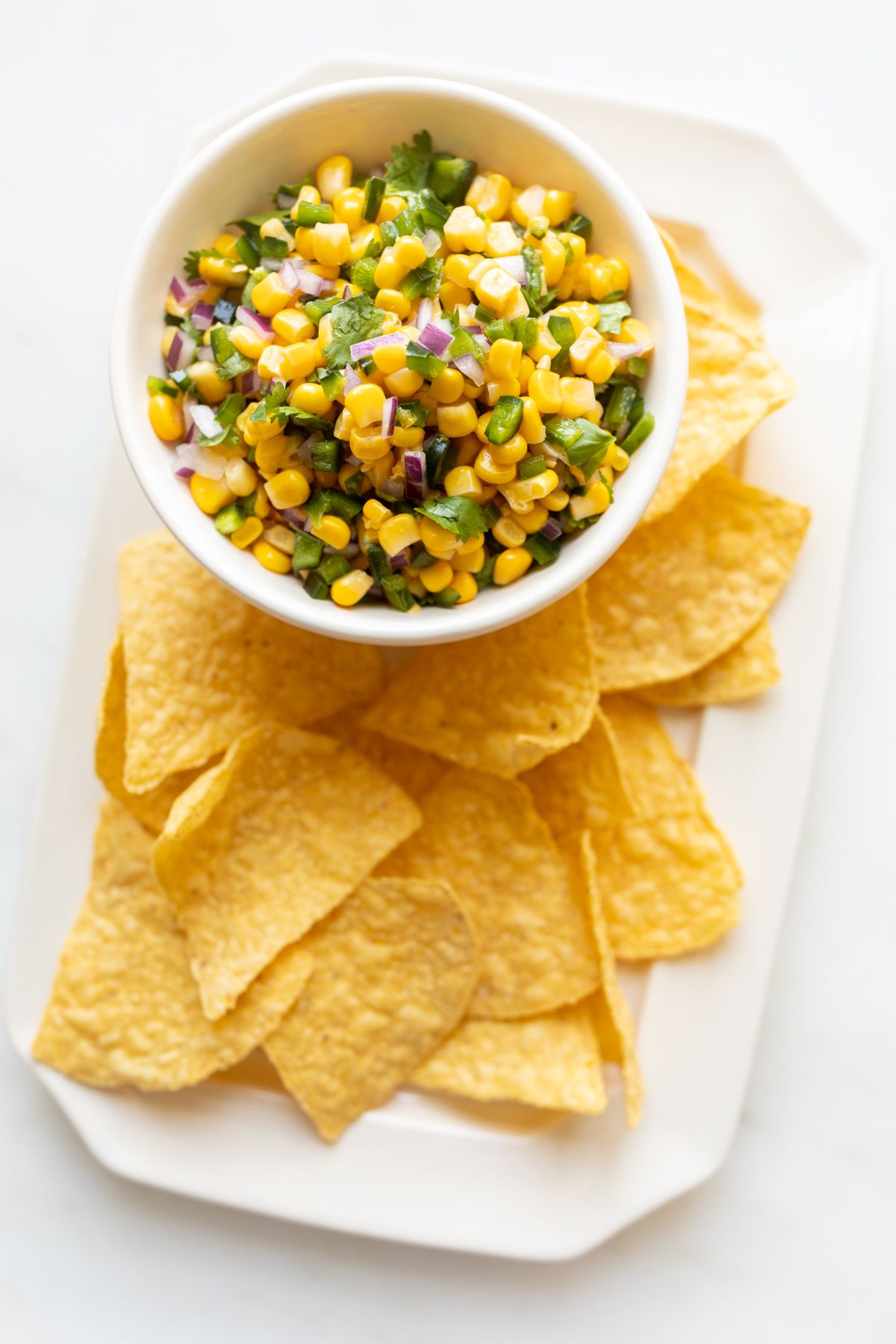 A chipotle corn salsa recipe in a white bowl, placed on a white platter full of corn tortilla chips