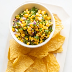 A chipotle corn salsa recipe in a white bowl, placed on a white platter full of corn tortilla chips