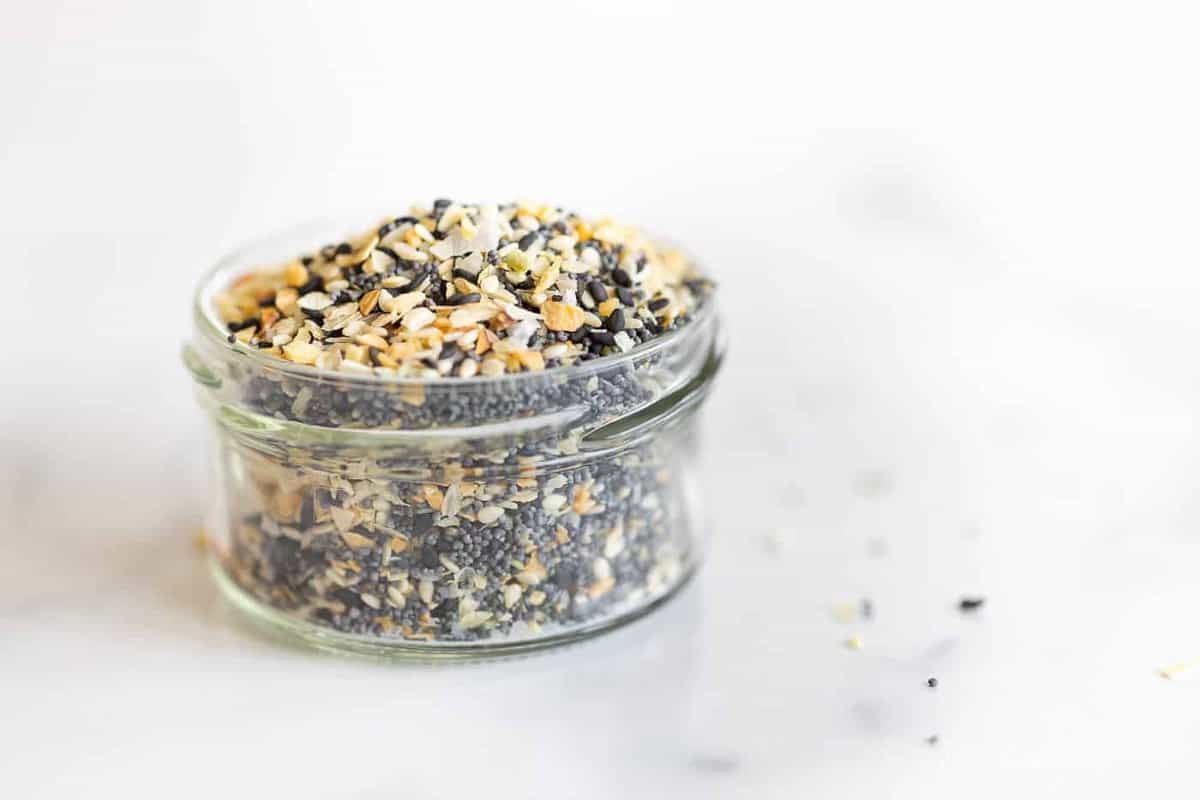 A clear glass jar on a marble surface, filled with everything bagel seasoning.