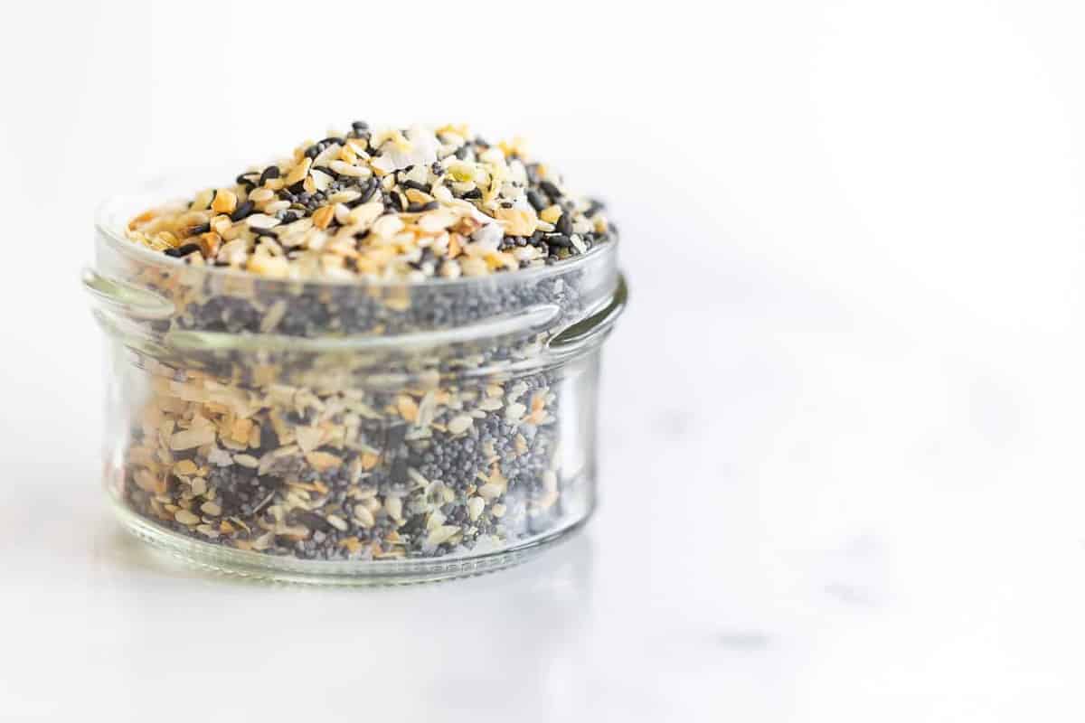 A clear glass jar on a marble surface, filled with everything bagel seasoning.