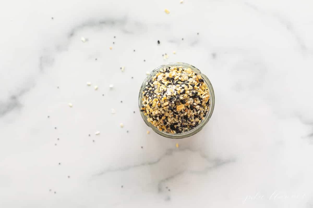 Everything bagel seasoning in a glass bowl and scattered on a marble surface.