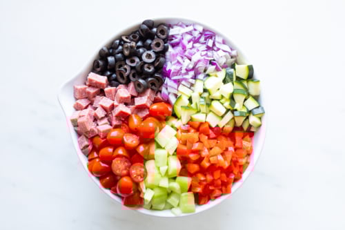 Ingredients for spaghetti salad in a white bowl on a marble countertop.
