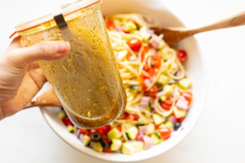 Spaghetti salad in a white bowl with wooden spoons for serving. A hand holds a jar of pasta salad dressing in the foreground.