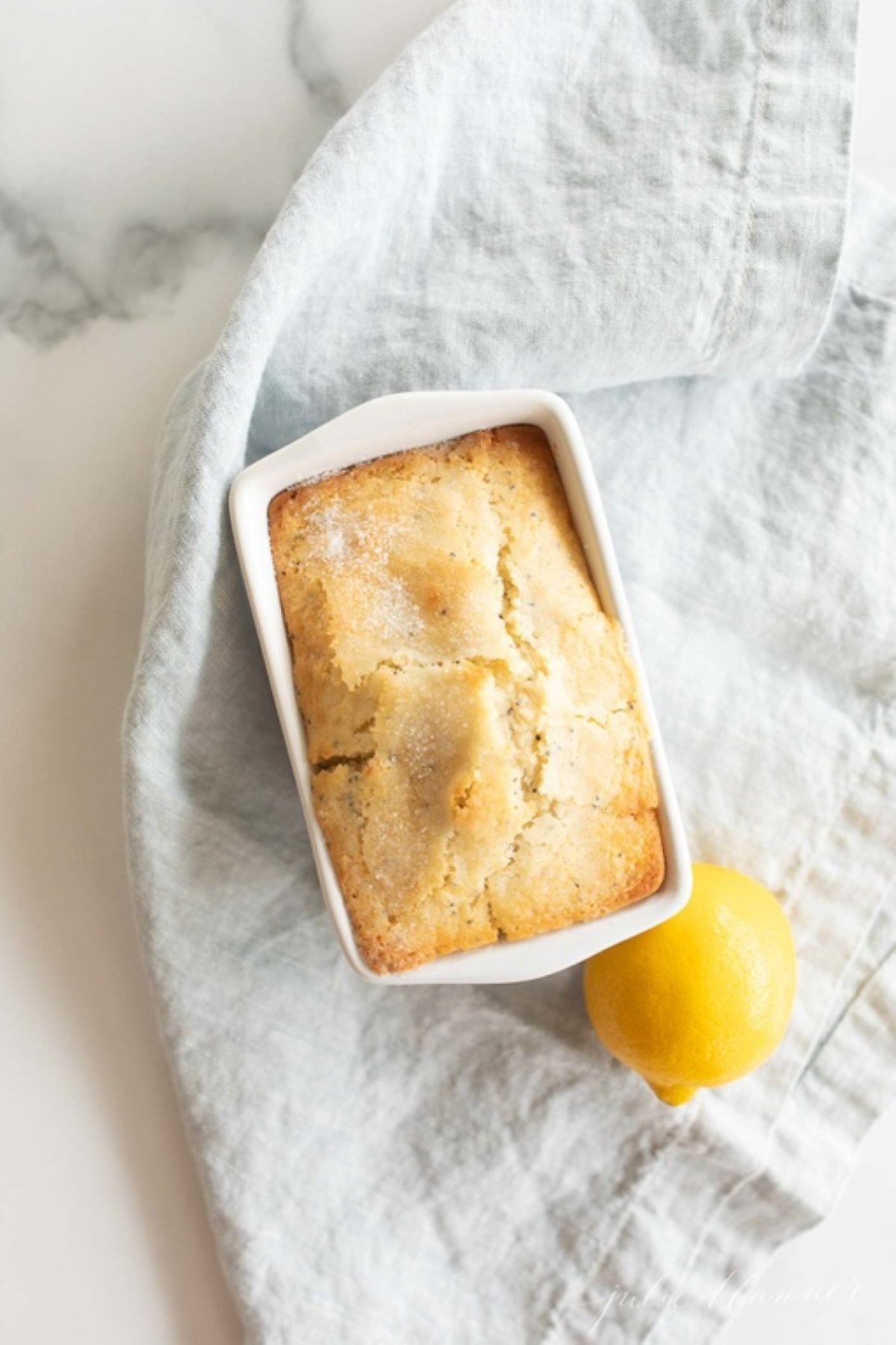 A small loaf of lemon poppy seed bread in a white ceramic loaf pan, with a lemon placed beside it.