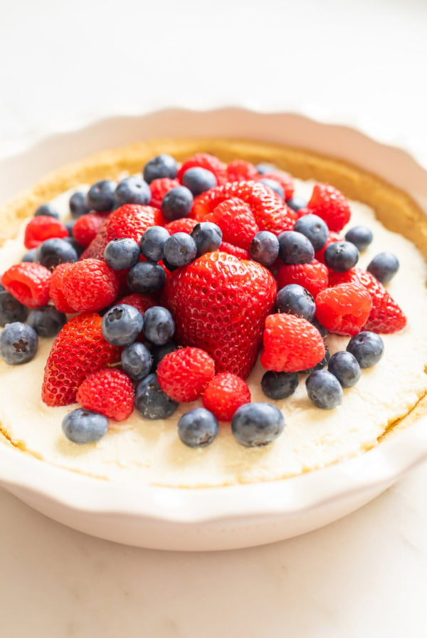 A no bake lemon pie cheesecake topped with fresh strawberries, blueberries, and raspberries in a white ceramic dish.