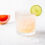 A paloma cocktail with a lime garnish and a slice off grapefruit in the background