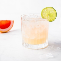 A paloma cocktail with a lime garnish and a slice off grapefruit in the background