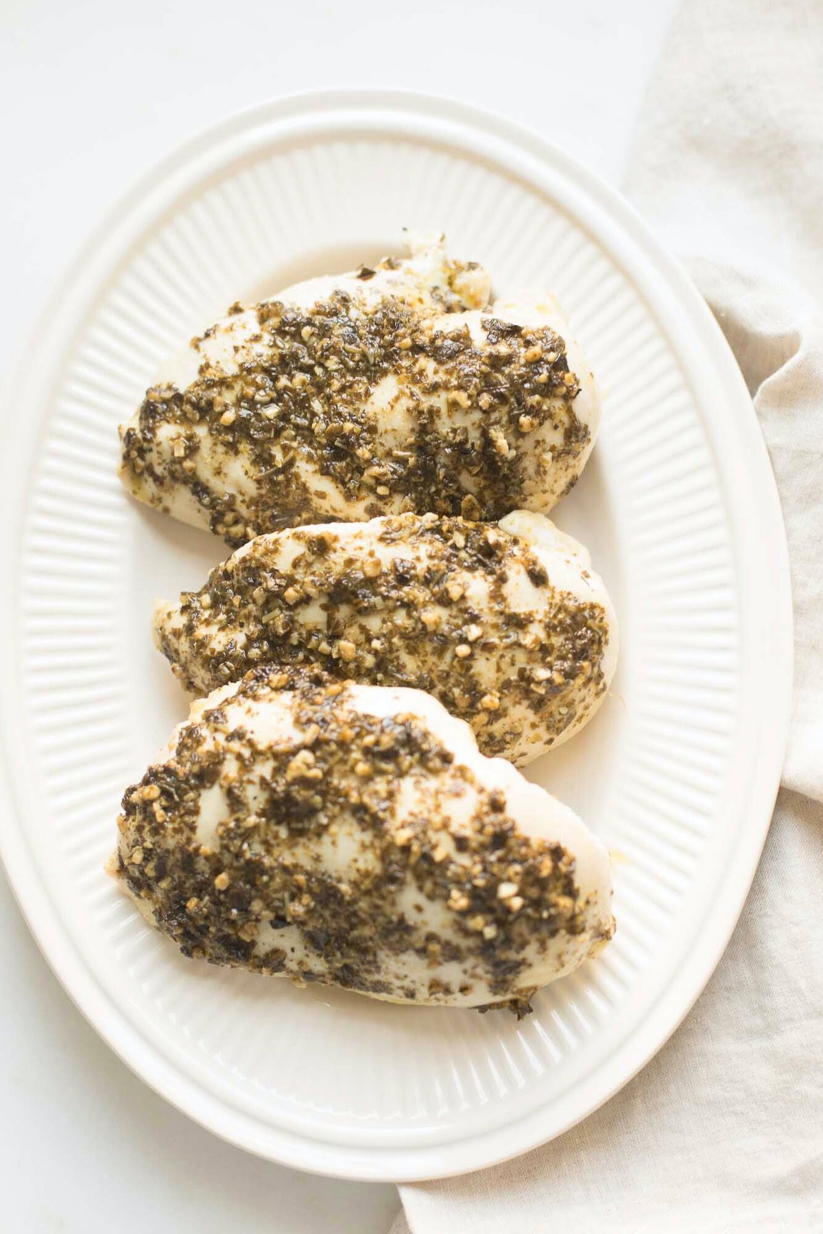 Three cooked chicken breasts coated with a herb and spice mixture, arranged on a white oval plate set against a light background—a delightful pesto chicken recipe.