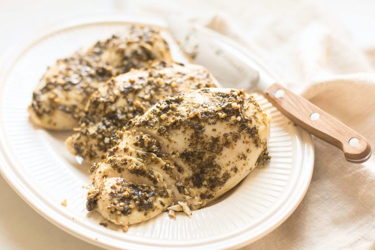 Sliced chicken breasts seasoned with herbs and spices, from a delightful pesto chicken recipe, served on a white plate with a knife in the background.