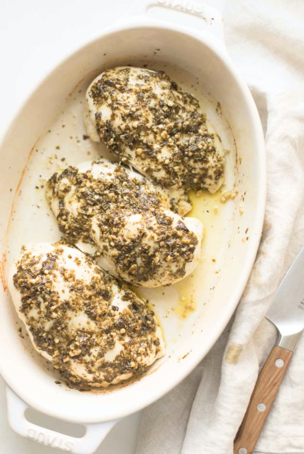 Three baked chicken breasts, seasoned with herbs and spices, bask in a white casserole dish. This succulent pesto chicken recipe is perfectly paired with a knife nearby for easy serving.