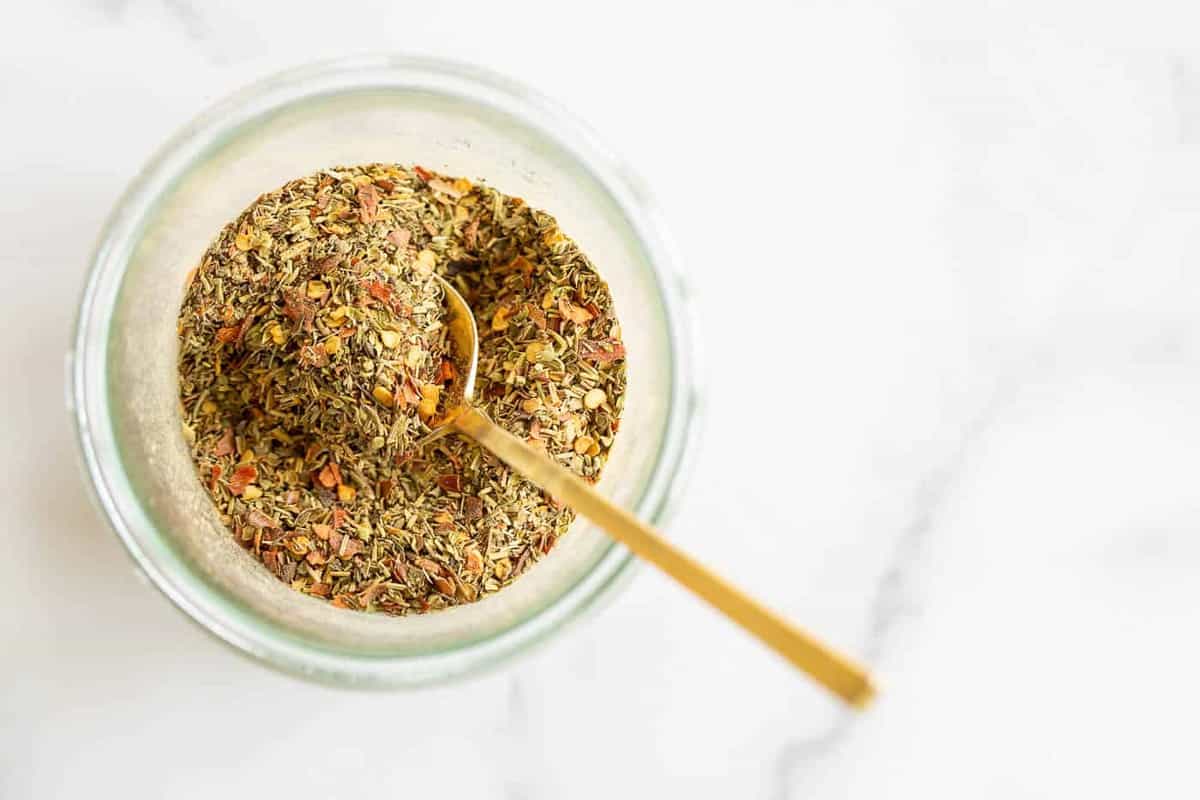 A clear glass jar of italian seasoning blend, gold spoon sticking out.