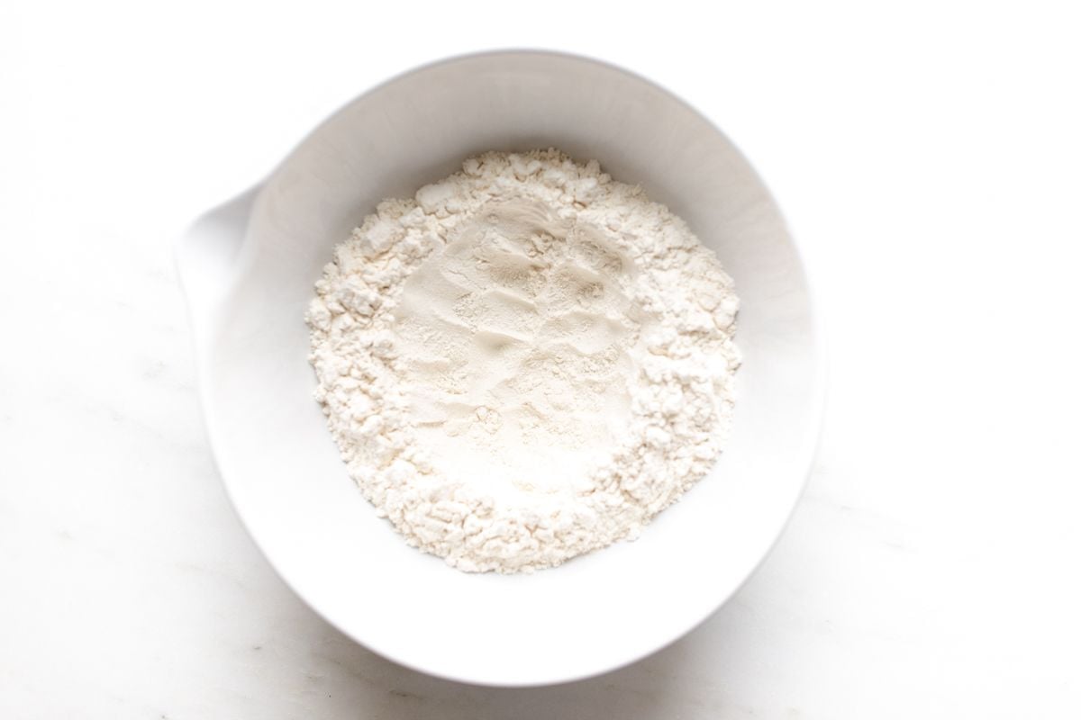 A white mixing bowl full of flour, with a well shaped into the center for a Belgian waffle recipe. 