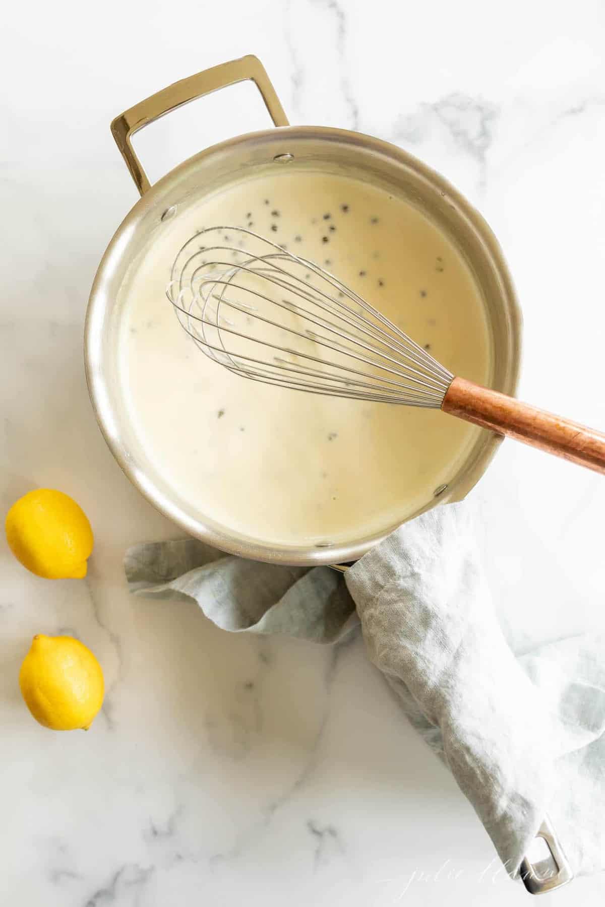 A pot full of white wine lemon caper sauce, with a whisk in the pot and lemons and linen napkins surrounding it. 