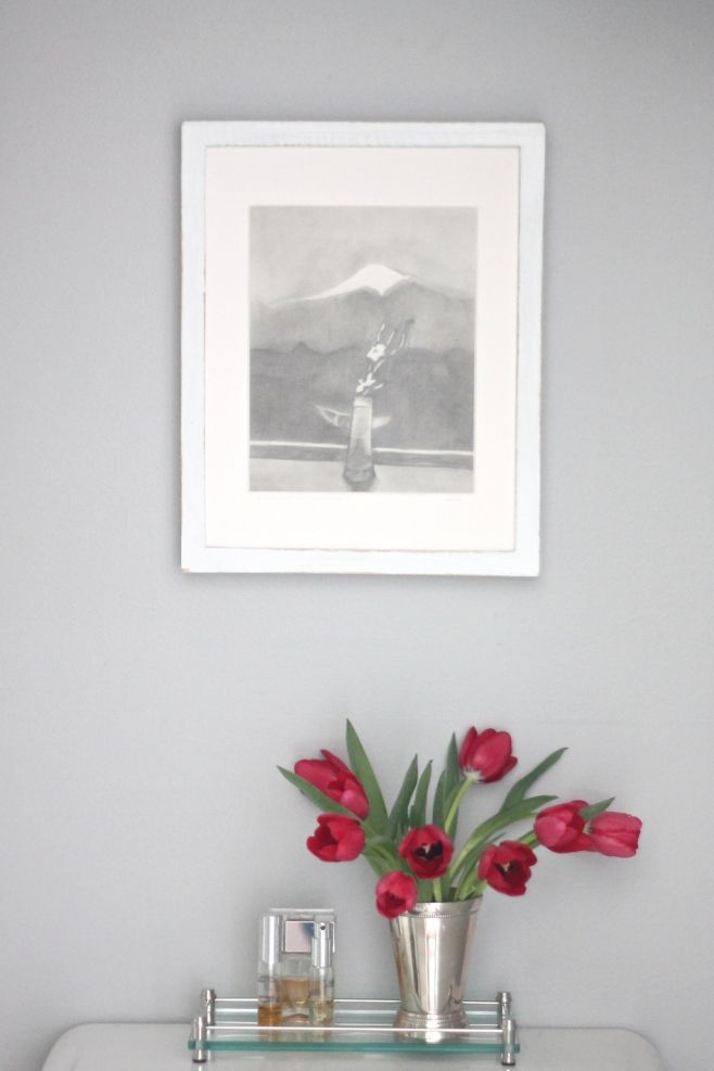 Decor over a toilet, including Knitting Needles wall paint, a vase of red tulips and framed art.