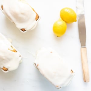 Three lemon-glazed pastries on a white surface next to two whole lemons and a spreading knife, ready for the lemon glaze icing.