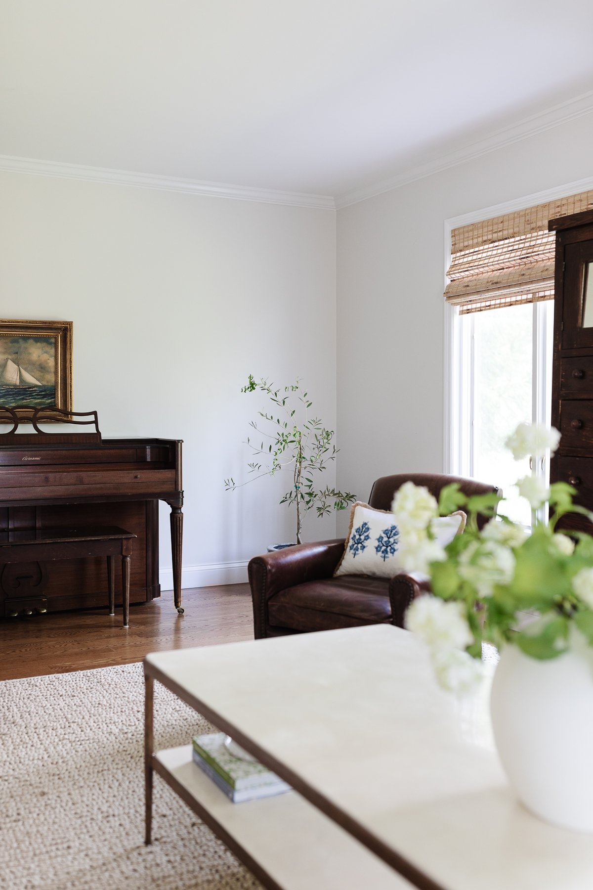 A well-lit living room with an upright piano, leather armchair, and a coffee table adorned with a vase of cut flowers. An easy care houseplant is in the corner.