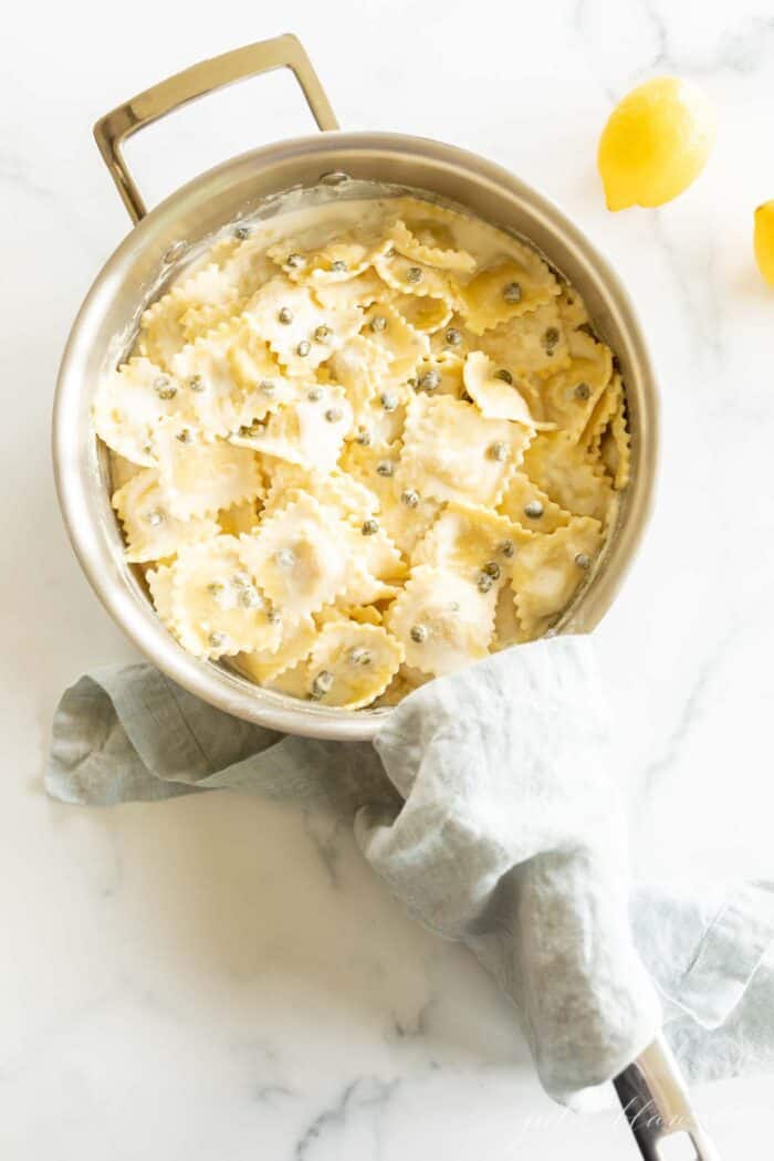 A silver pot filled with ravioli in a white wine lemon caper sauce on a marble surface.