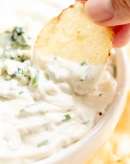 blue cheese dip in a white bowl, with fingers gripping a chip to dip into it.