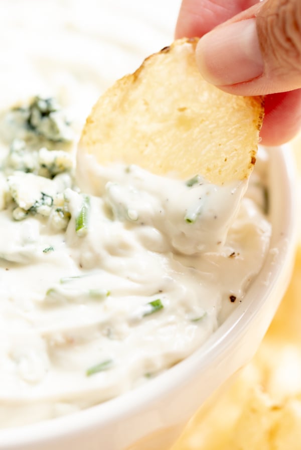 blue cheese dip in a white bowl, with fingers gripping a chip to dip into it.