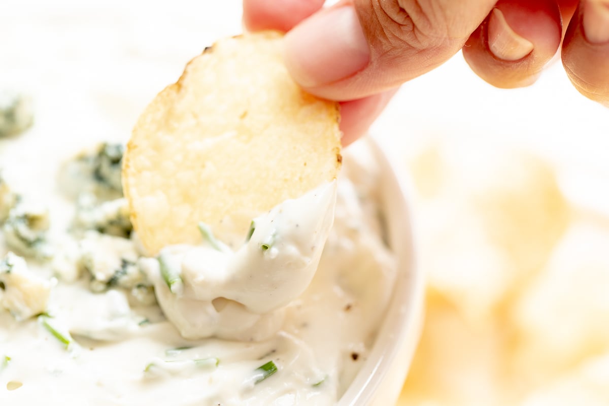 blue cheese dip in a white bowl, with fingers gripping a chip to dip into it.