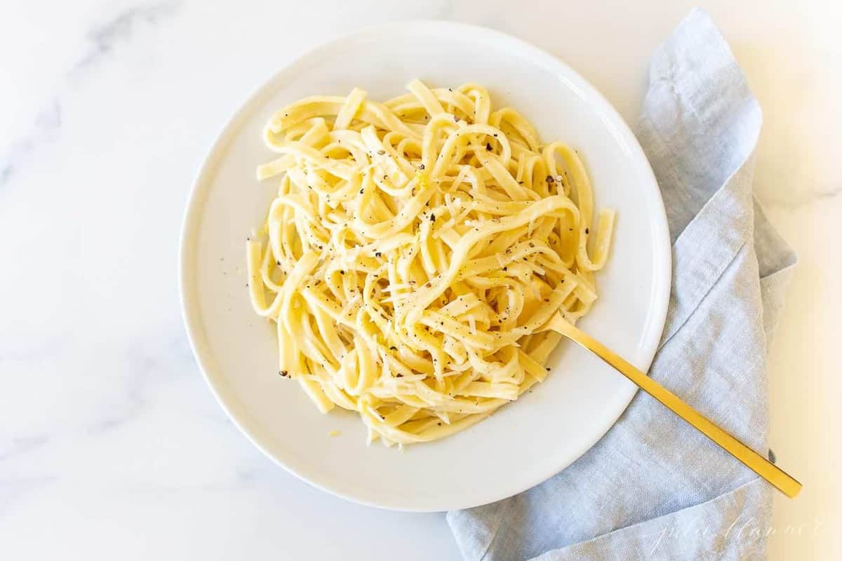 A white plate with a bowl full of lemon pepper pasta, gold fork to the side.